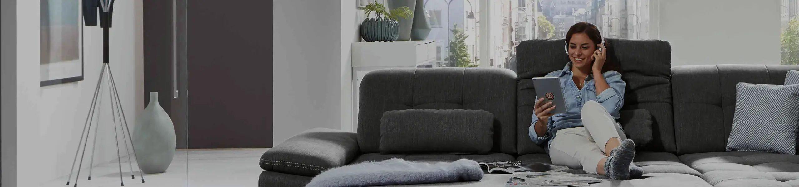 Frau sitzt entspannt mit Tablet auf einem großen grauen Ecksofa. Komfortables Sofa mit Relaxfunktion und moderner Einrichtung, ideal für gemütliche Stunden im Wohnzimmer.